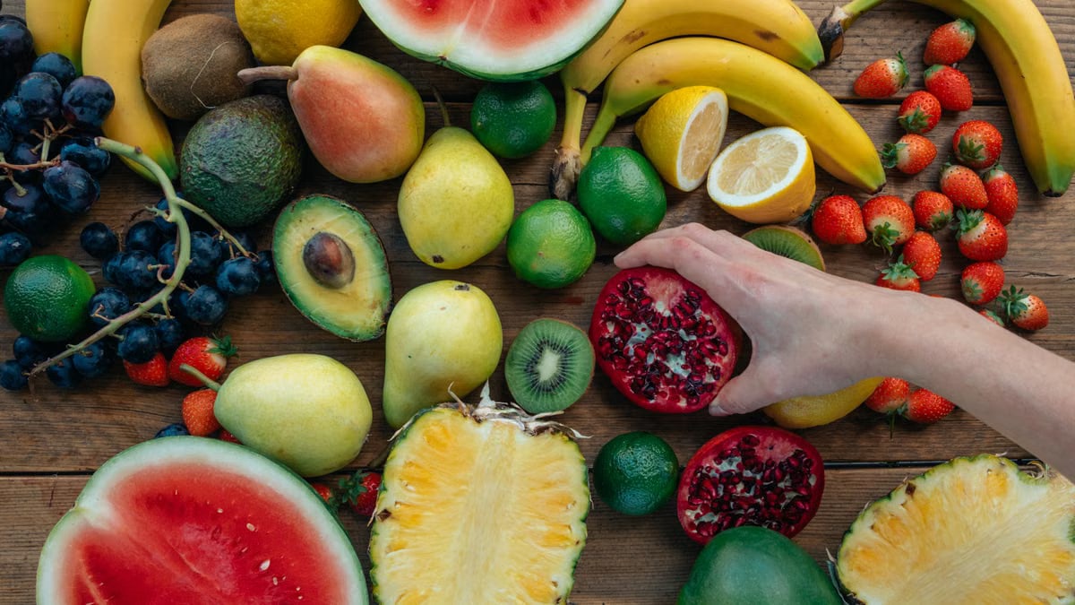 Colorful assortment of fresh fruits on wooden surface with a hand holding a pomegranate half.